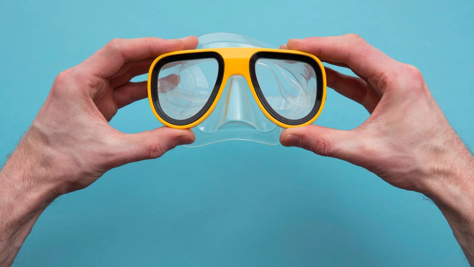 A pair of hands holding a dive mask up against a plain background