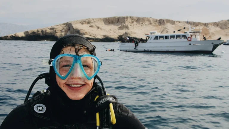 A diver in full scuba equipment smiling at the surface with a boat behind