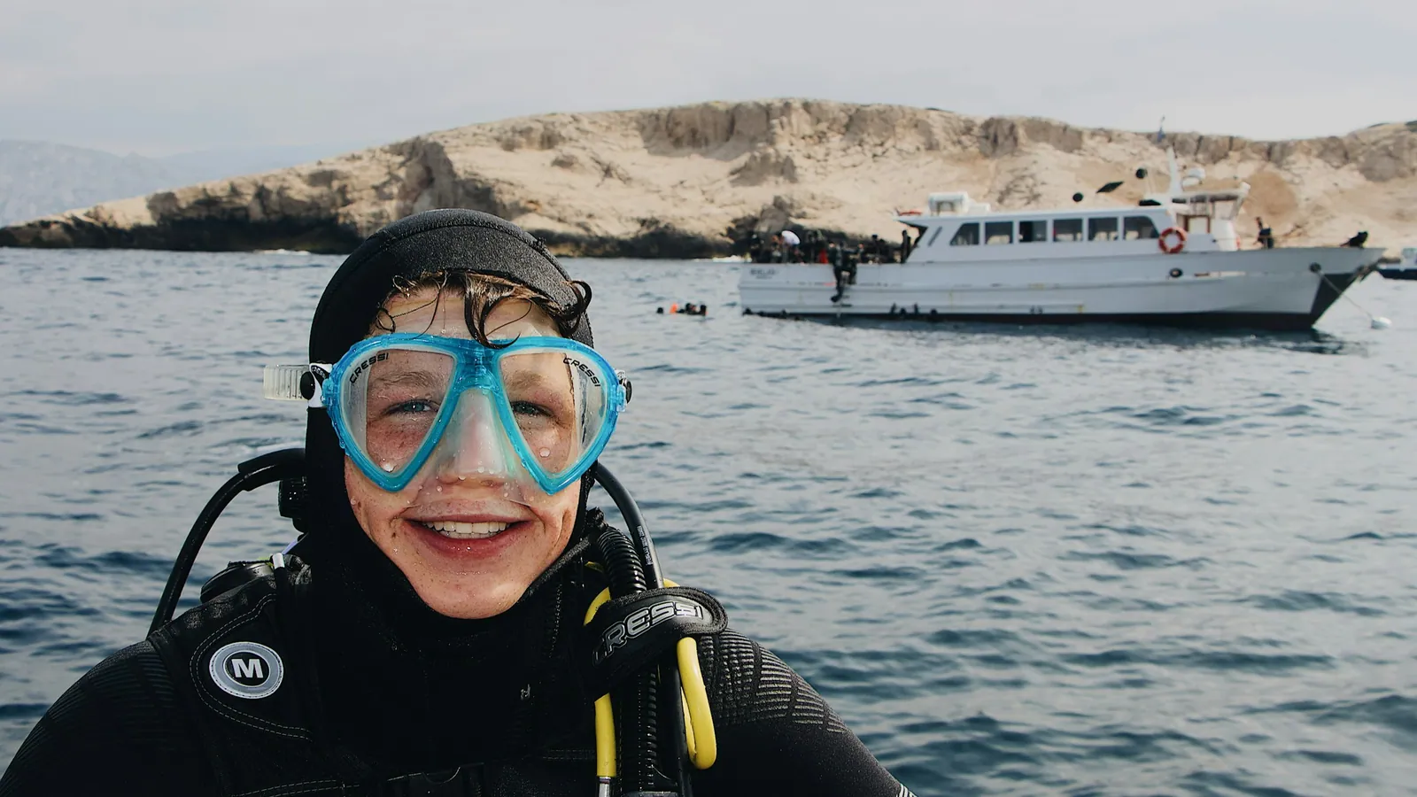 A diver in full scuba equipment smiling at the surface with a boat behind