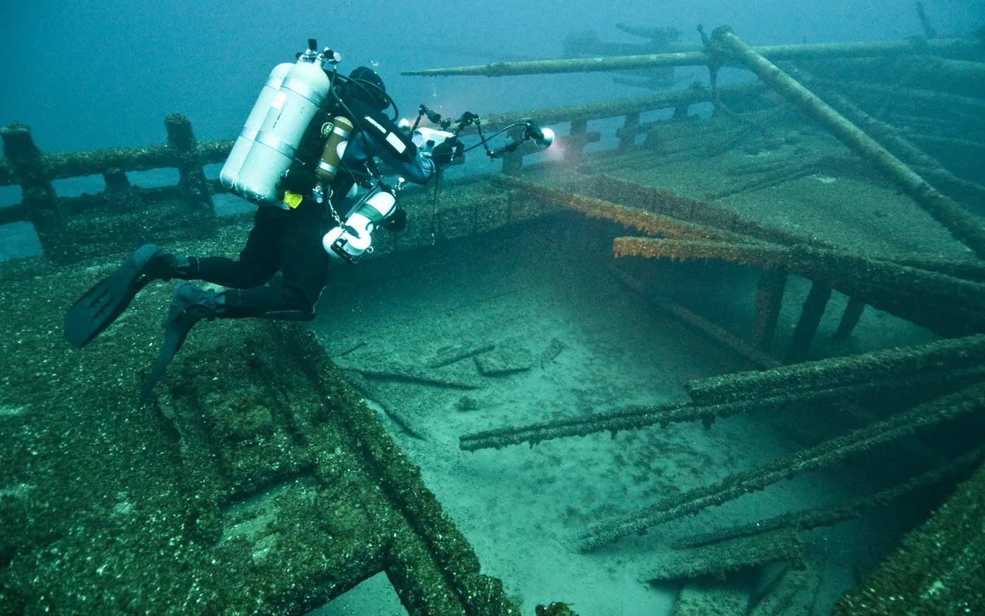 A scuba diver with twin cylinders and lights above a collapsed wreck