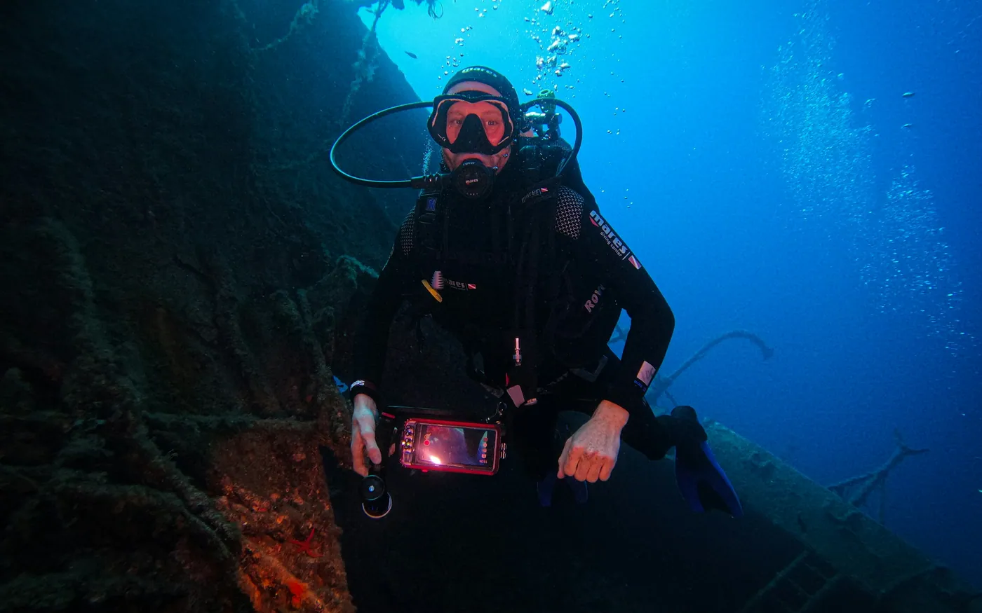 A scuba diver holding a camera alongside a wreck in blue water