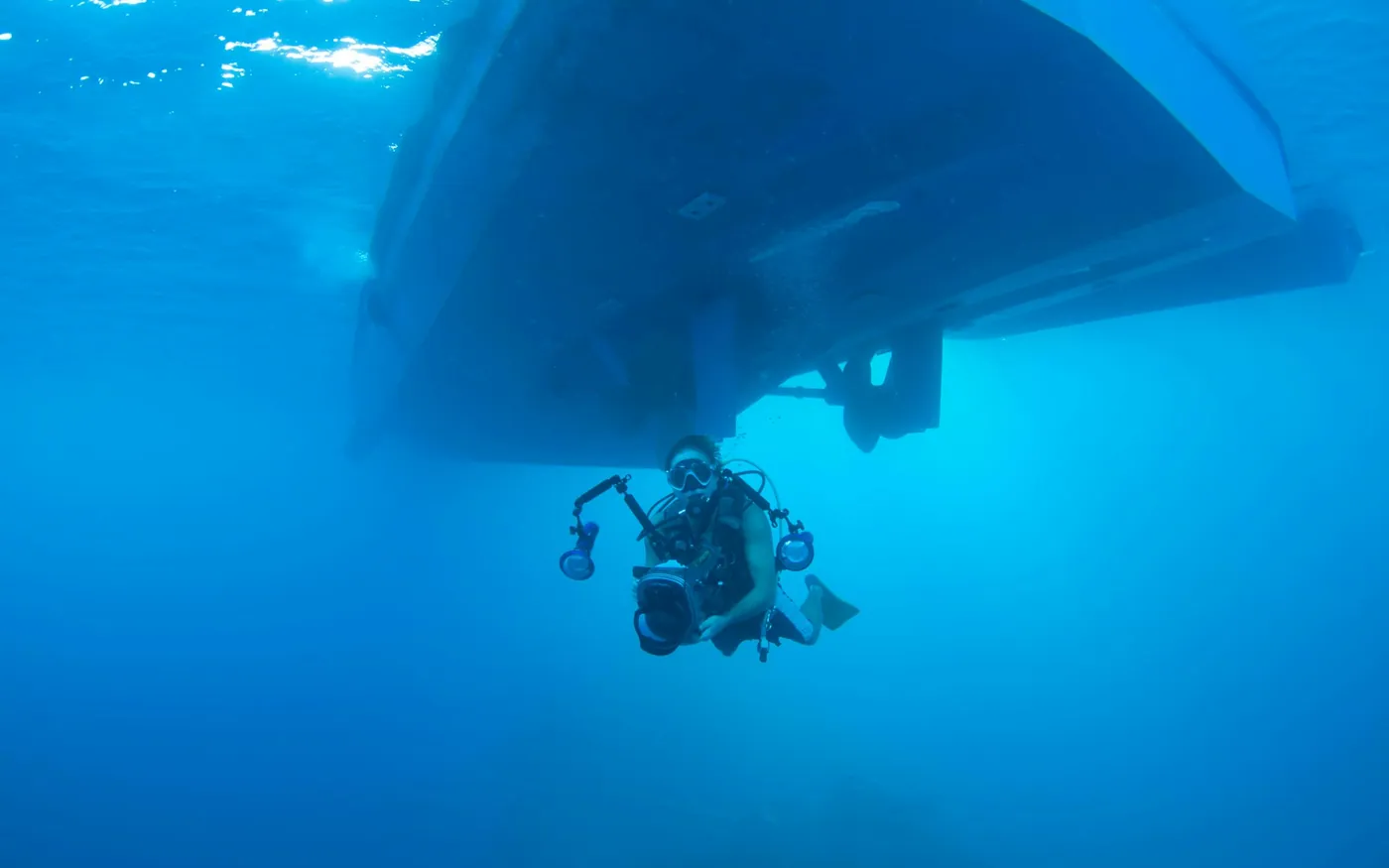 A scuba diver carrying a camera rig beneath the hull of a boat