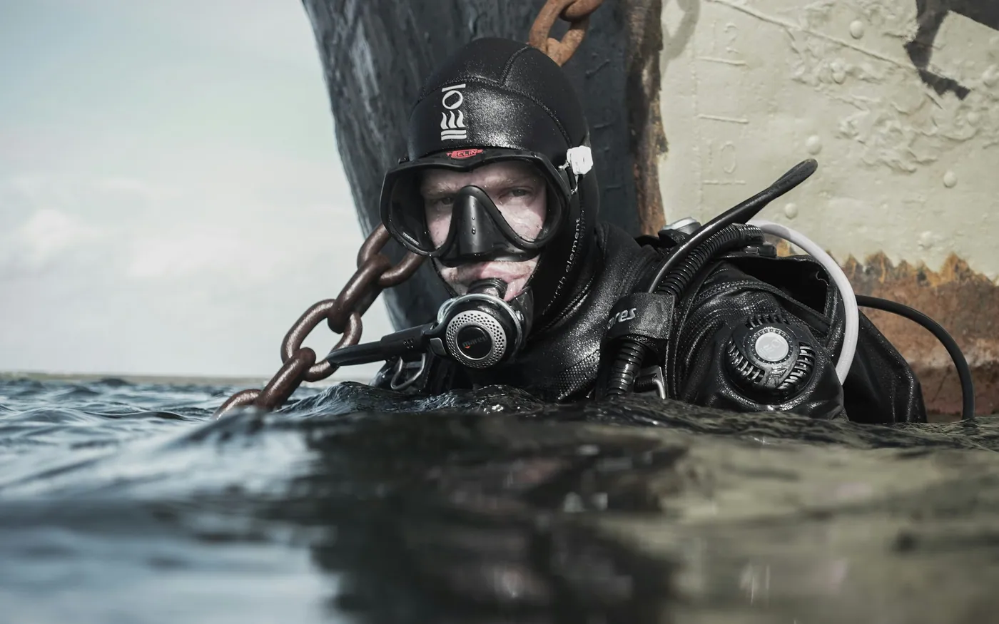 A diver in a hood at the surface holding a chain in cold grey water