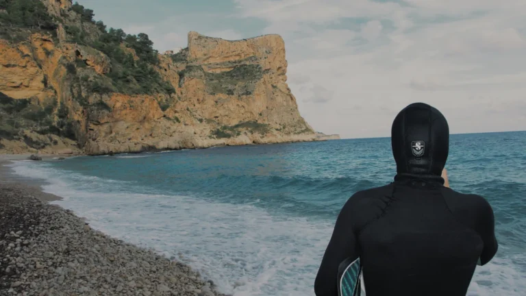 A diver in a hooded wetsuit standing on a pebble beach looking out at grey water