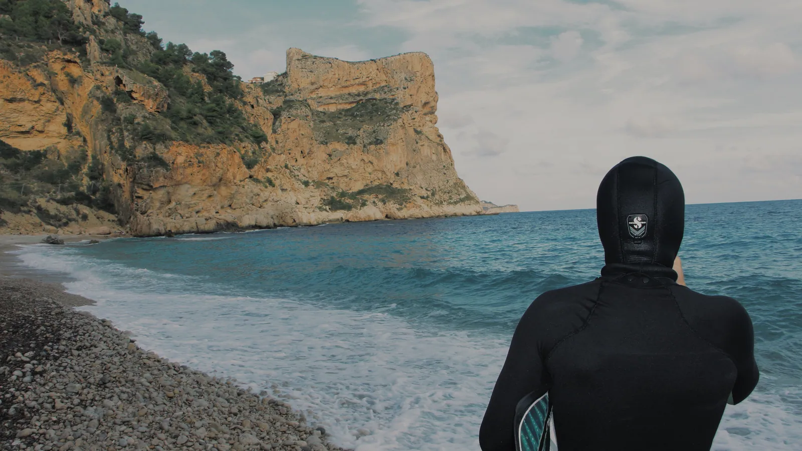 A diver in a hooded wetsuit standing on a pebble beach looking out at grey water