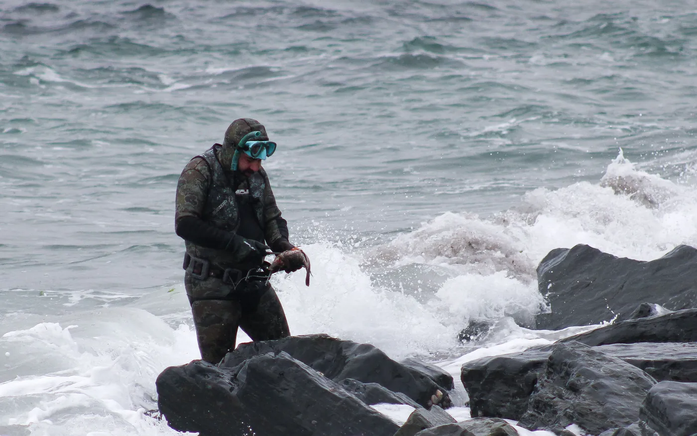A diver in a hooded drysuit climbing over rocks in rough grey water on a cold-water shore dive
