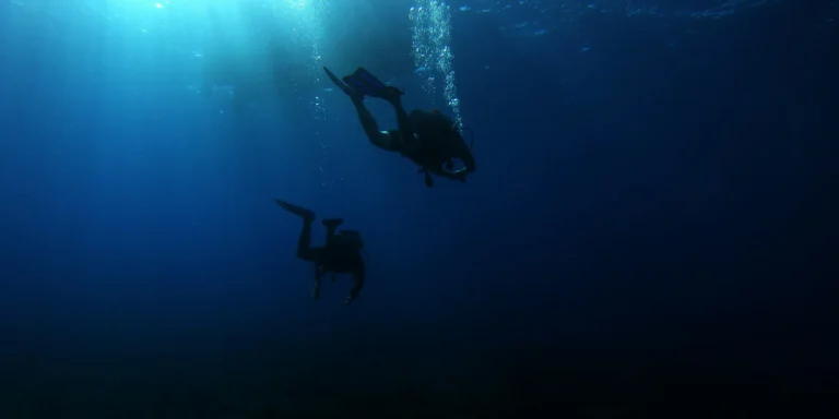 Two scuba divers with cylinders and rising bubble trails descending together in deep blue water beneath a boat