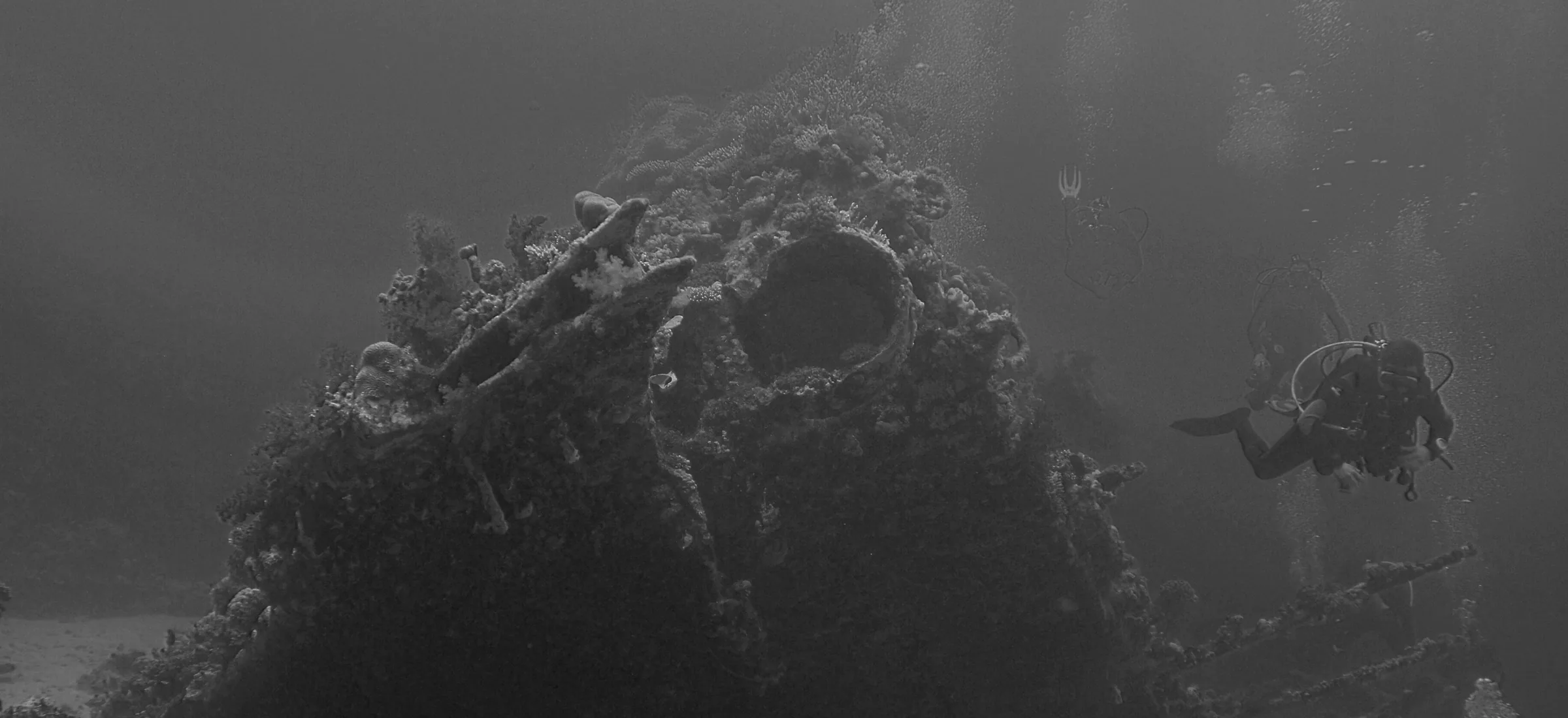 Scuba divers with cylinders and regulators exploring a shipwreck in grey-green, low-visibility water, exhaust bubbles rising