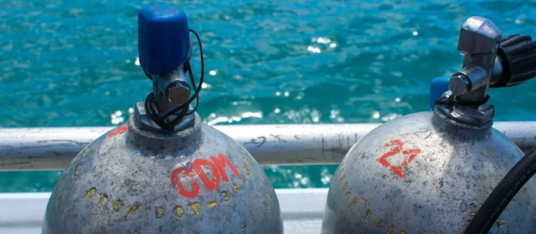 The crowns and valves of two scuba cylinders standing on a boat rail, with the sea behind