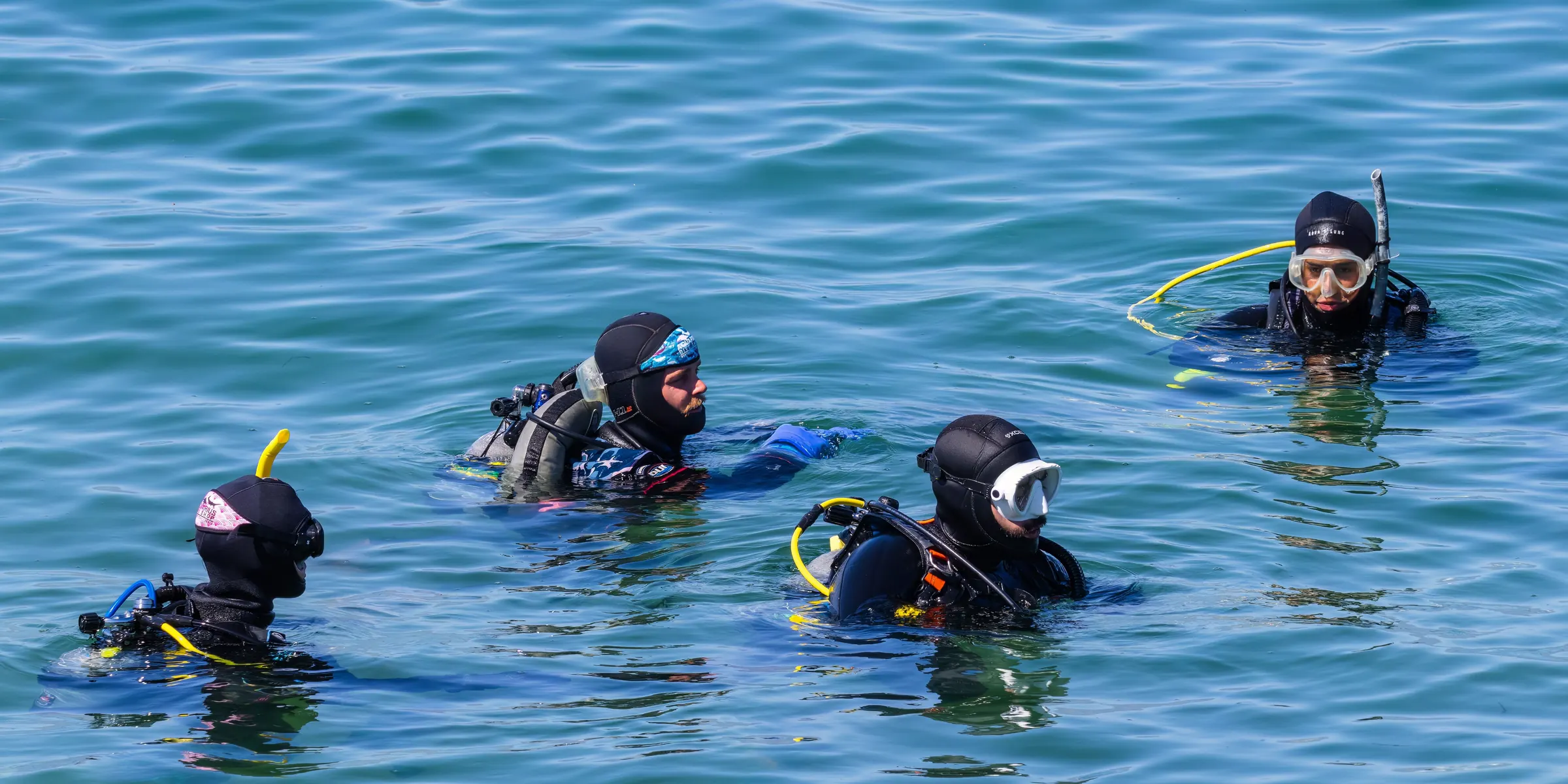 Four scuba divers in hooded wetsuits with cylinders and regulators, waiting at the surface in green water before descending