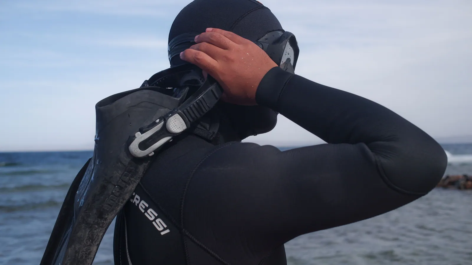 A diver in a hooded wetsuit adjusting a mask at the water's edge