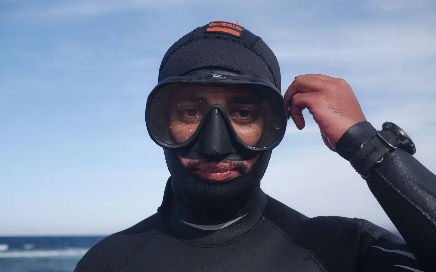 A diver in a wetsuit hood adjusting a mask strap before a dive