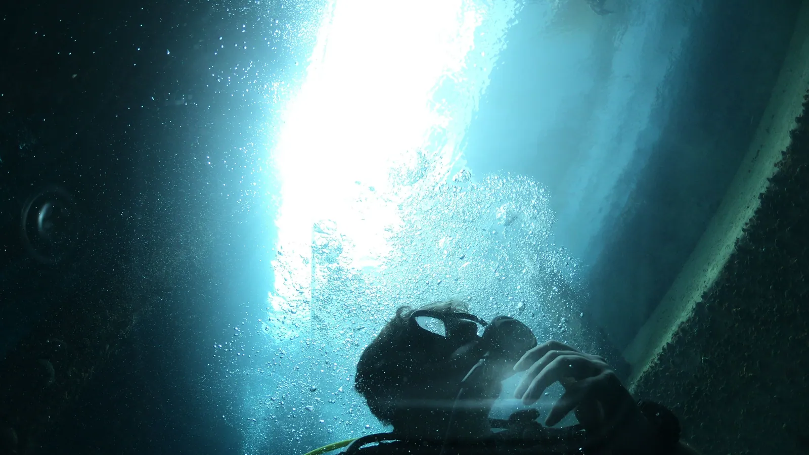 A scuba diver below a boat hull with sunlight breaking through the surface