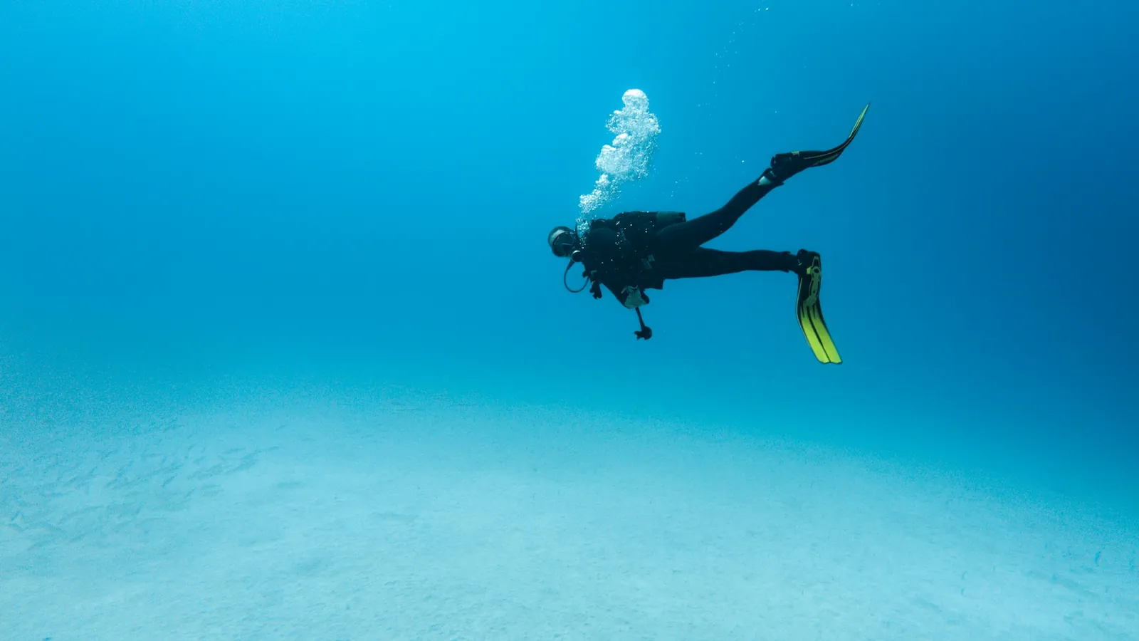 A scuba diver hovering horizontally in mid-water above a sandy bottom