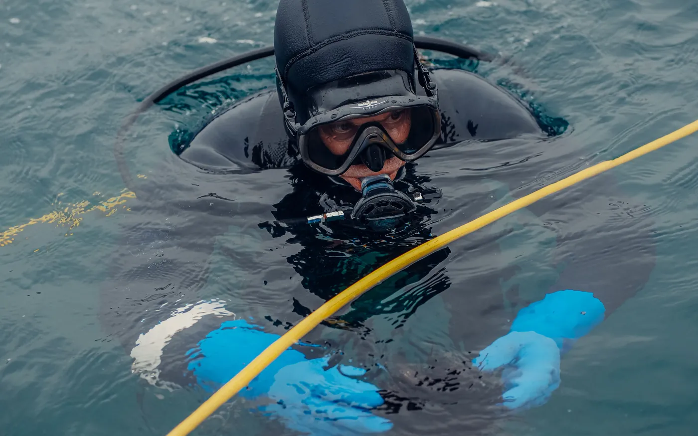 A diver in a hooded wetsuit and mask at the surface in cold green water