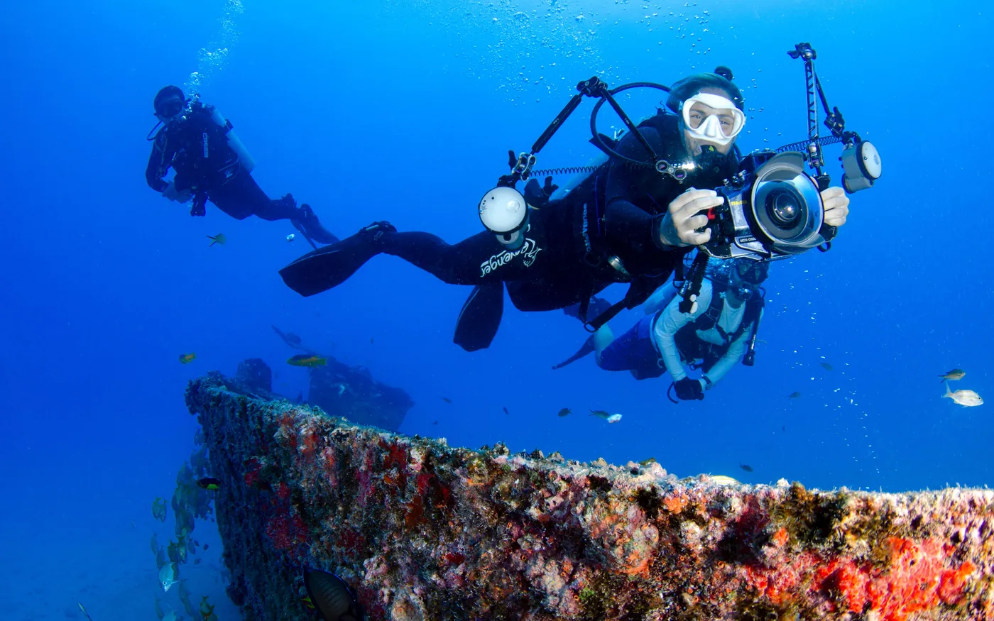 A scuba diver carrying an underwater camera rig above an encrusted wreck