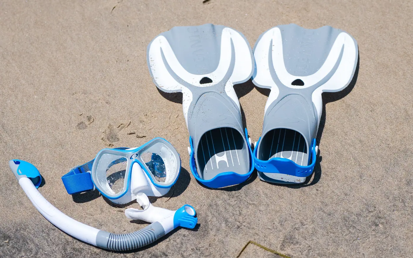 A dive mask, snorkel and a pair of open-heel fins laid out on sand