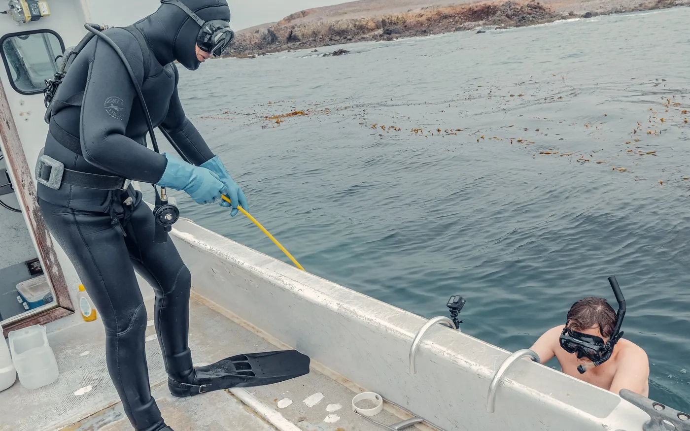 A diver on a boat deck handling a hose while another diver waits in the water