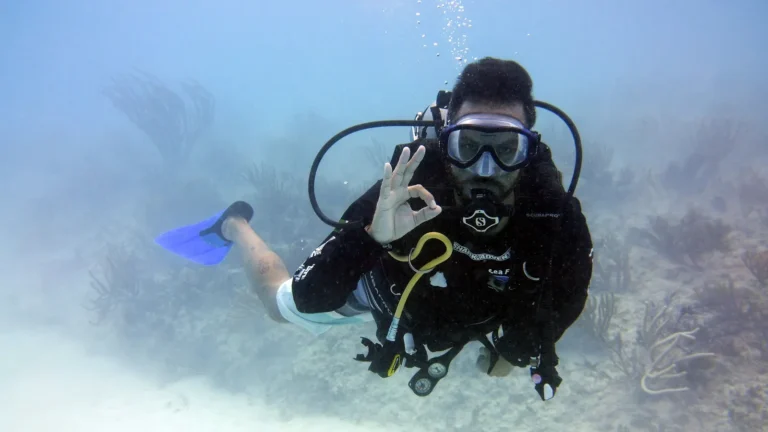 A certified scuba diver in full gear signalling OK underwater over a sandy seabed