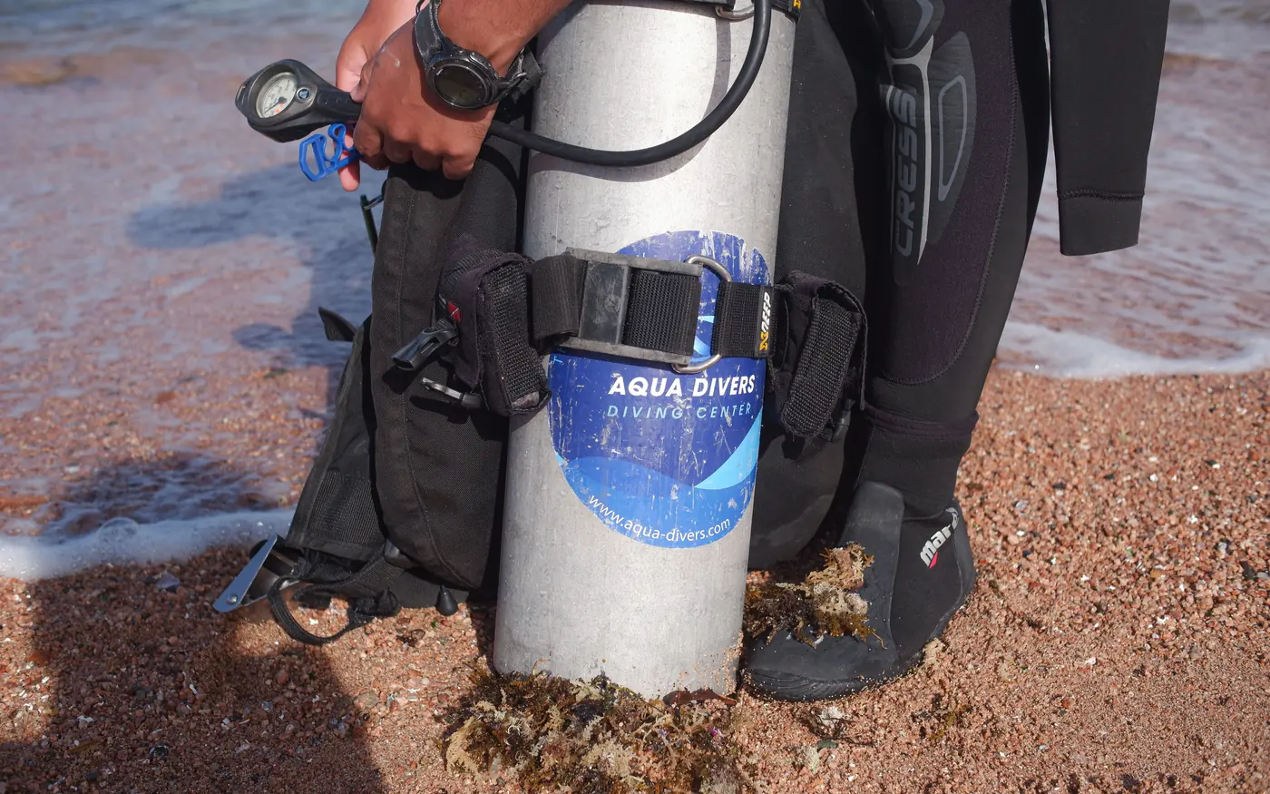 A scuba cylinder rigged with a BCD and regulator standing on the shore beside a diver