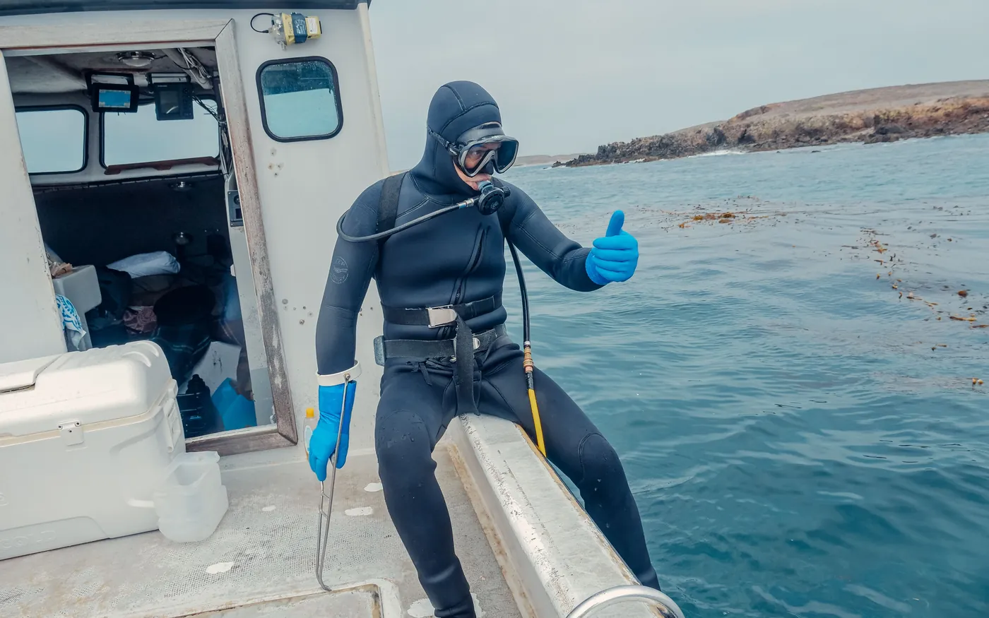 A scuba diver in a hooded wetsuit giving a thumbs up on the deck of a dive boat