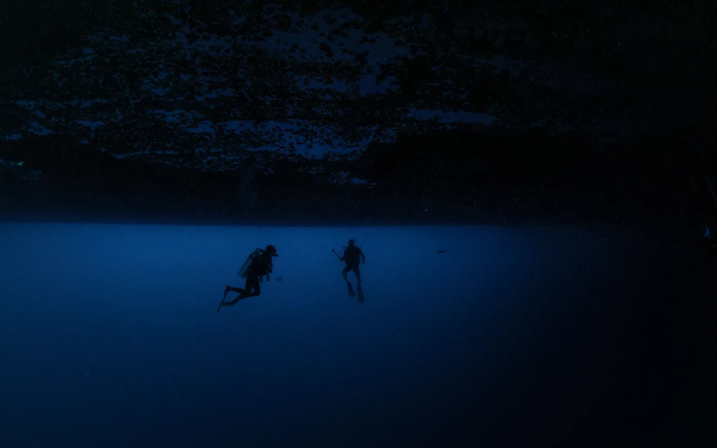 Two scuba divers silhouetted against a wall in very dark water