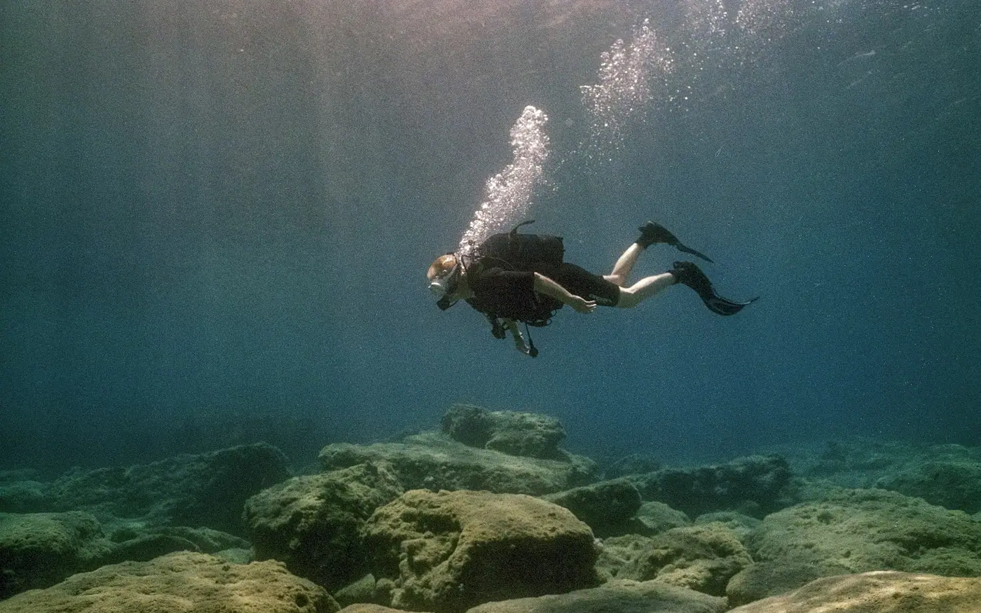 A scuba diver moving slowly above a rocky bottom in shafts of light
